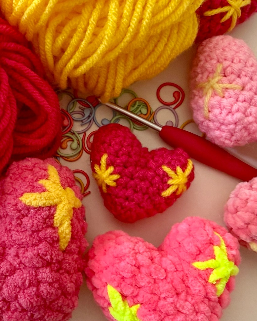 Crocheted sparkle heart emoji surrounded by yarn, a crochet hook, stitch markers, and alternate versions of the finished heart in different yarns.