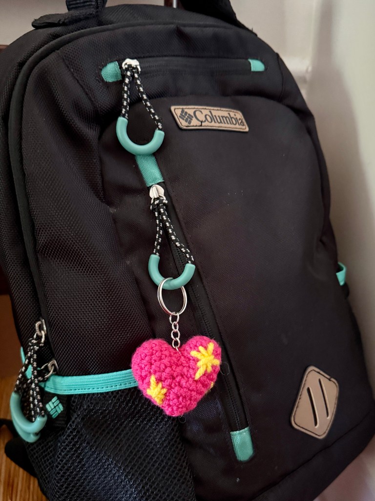 Close-up of a crocheted pink sparkle heart emoji keychain attached to a backpack, showing the finished project in use as a bag charm.